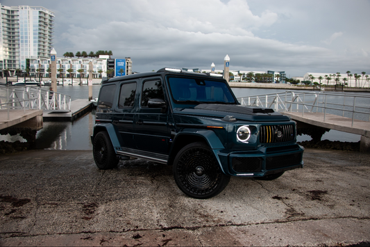 A dark metallic blue Mercedes-Benz G-Class SUV parked outdoors near a waterfront pier, with buildings and water visible in the background under an overcast sky.