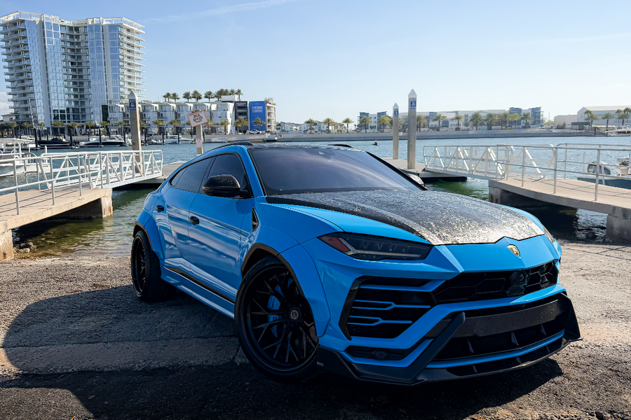 Blue Lamborghini Urus parked by a waterfront with buildings in the background