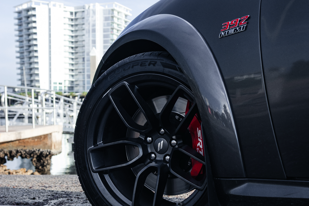 Close-up of a black car wheel with a visible brand logo on a road with buildings in the background.