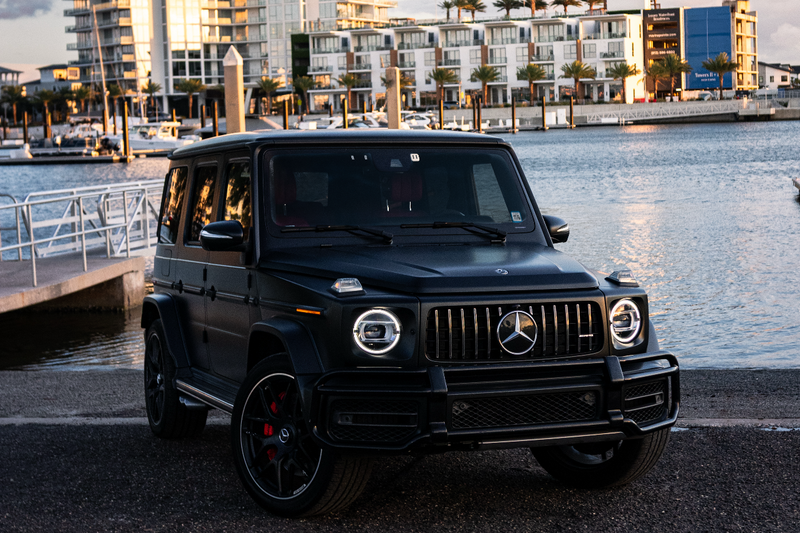 Black Mercedes-Benz G-Class SUV parked by a waterfront with buildings in the background