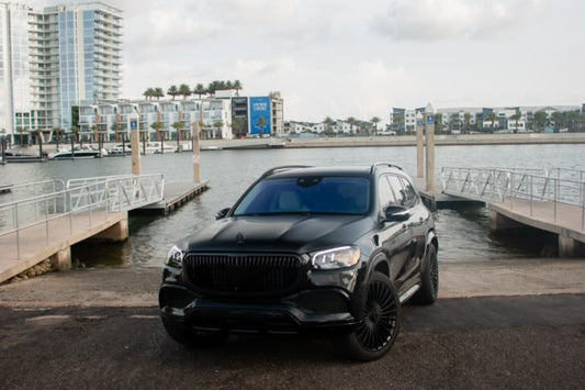 A black luxury SUV parked on a paved dock next to water, with a marina and modern buildings visible in the background under a cloudy sky.